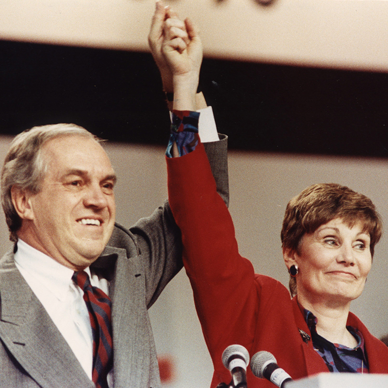 Ed Broadbent and Audrey Mclaughlin behind a podium with their hands raised together.